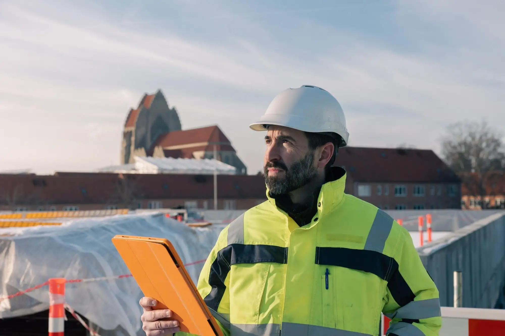 Byggledare med hjälm och orange skrivplatta framför en byggplats