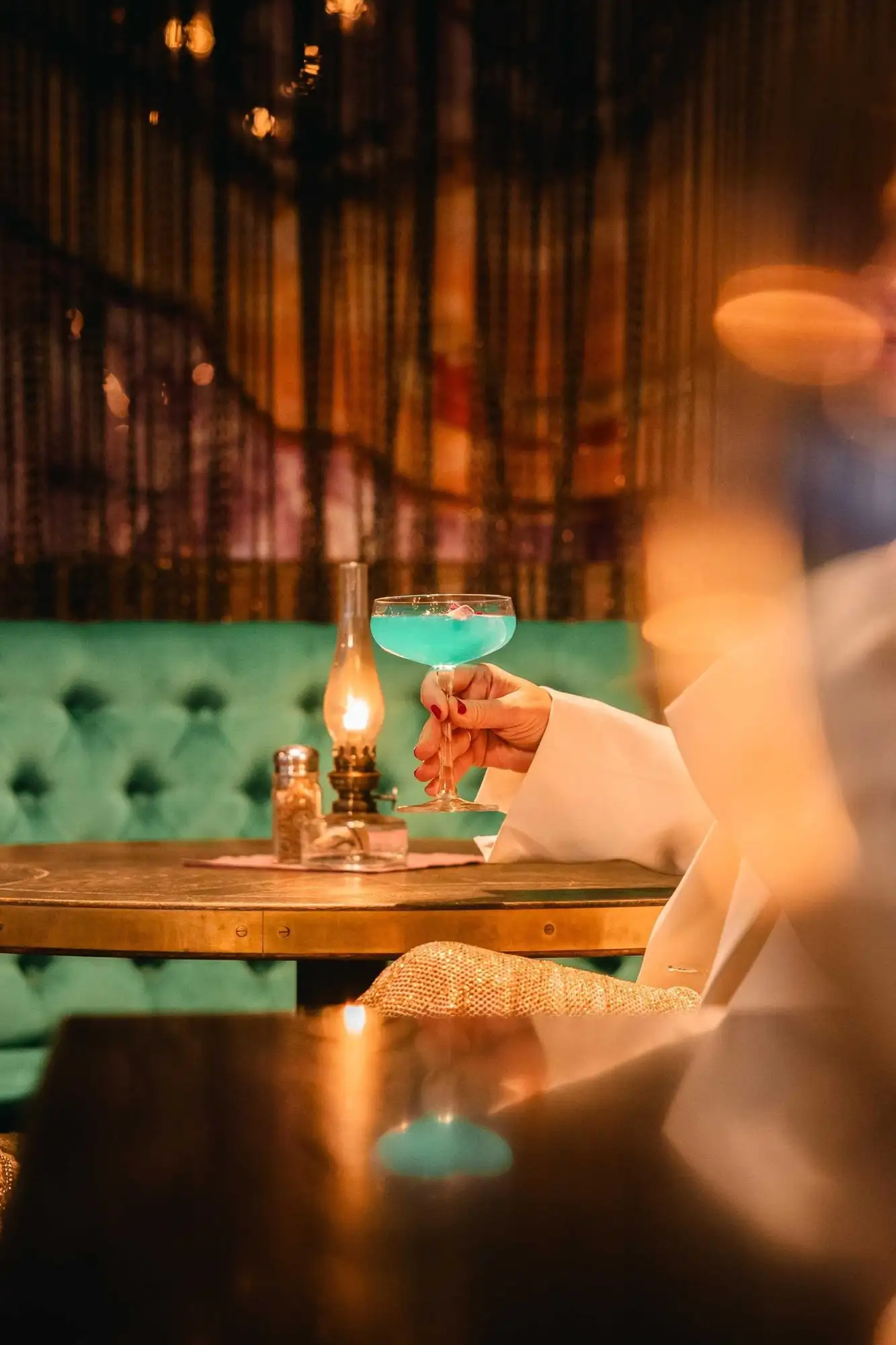 Hand med en blå cocktail i en barmiljö med grön soffa och levande ljus