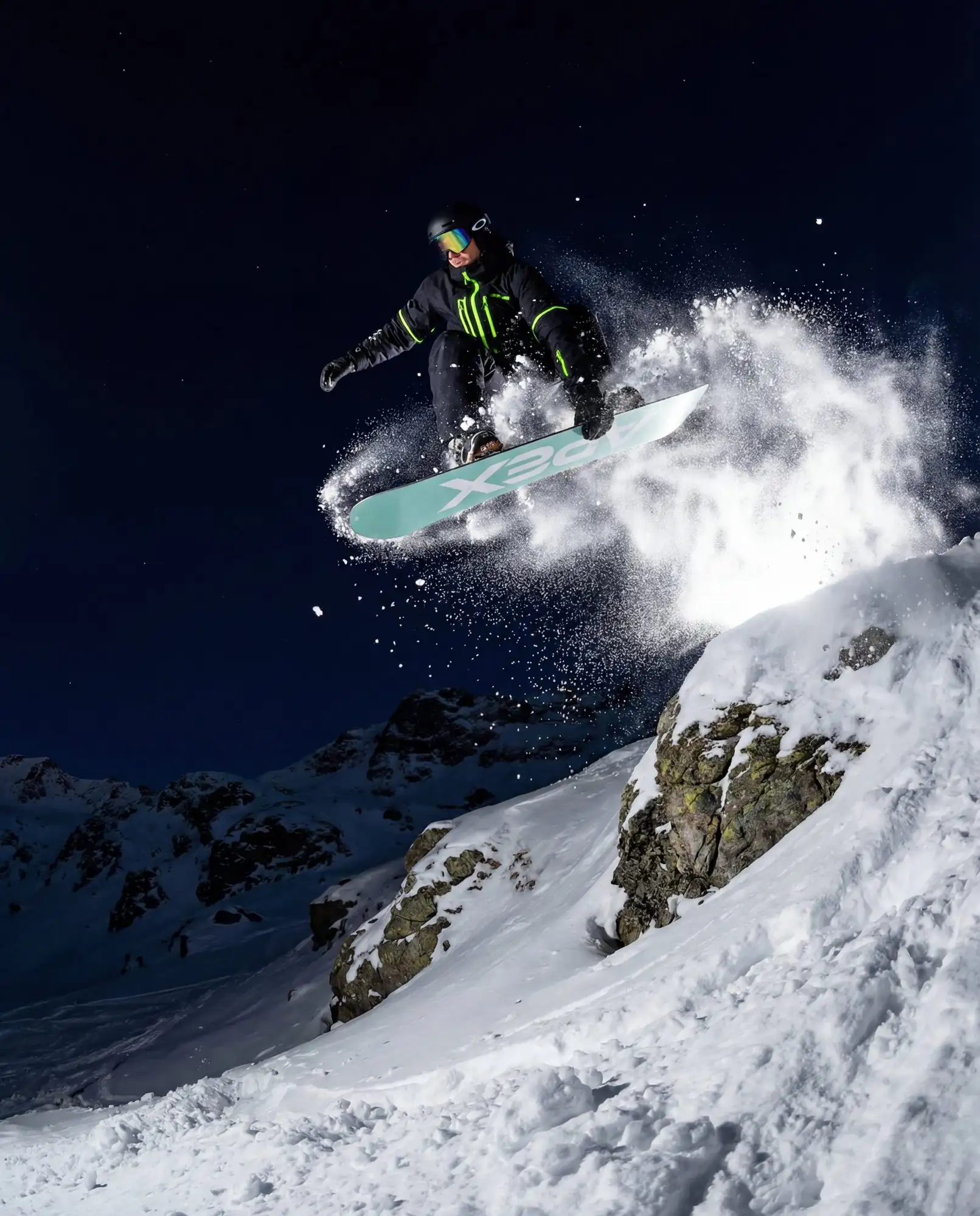 Snowboardåkare hoppar över en bergskam i nattljus med snöpuder i luften